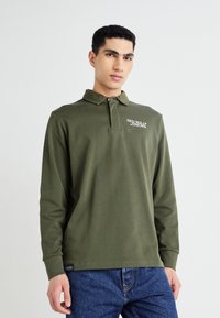 Polo majica z dolgimi rokavi v temno zeleni barvi iz bombaža s ovratnikom, ki ima bel logo "RED BULL RACING", kombinirana z modrimi kavbojkami.