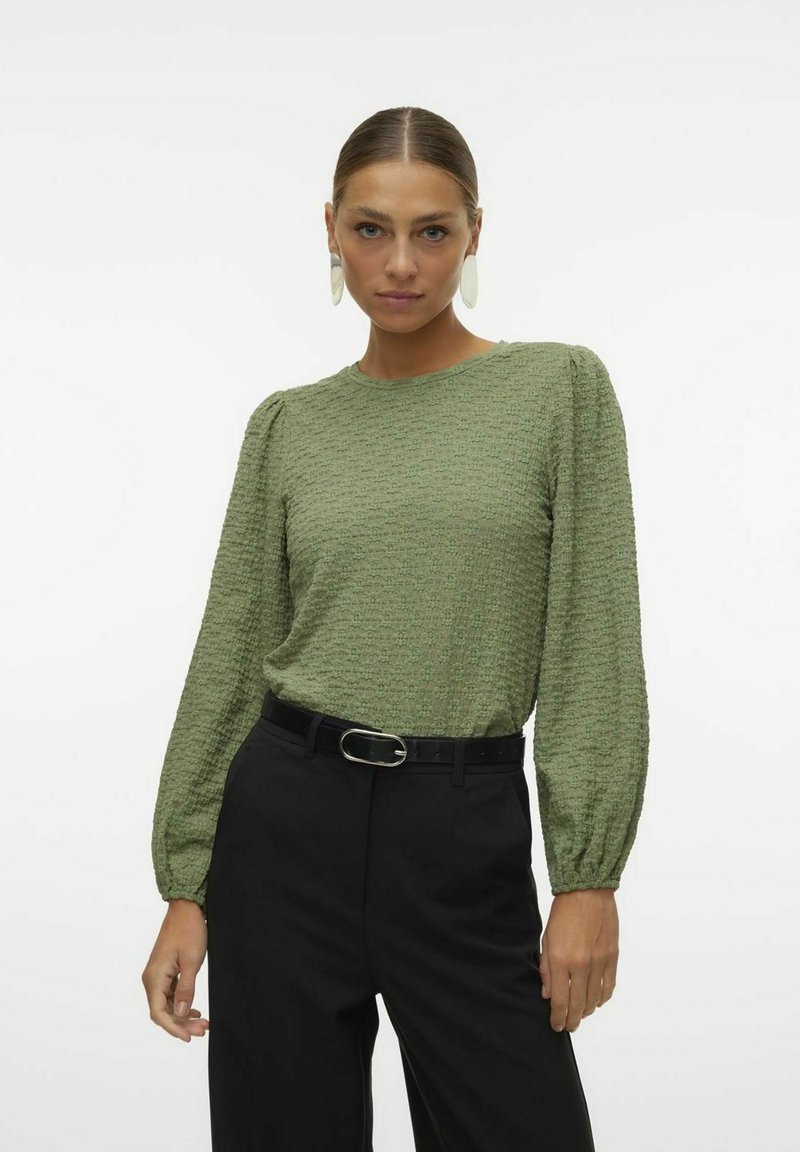 Blusa lunga a maniche lunghe verde texturizzata con spalle a palloncino, abbinata a pantaloni neri a vita alta e una cintura nera con fibbia argentata.