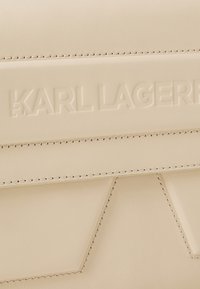Krem torba od kože s urezanim logotipom "KARL LAGERFELD", detaljnim šavovima i geometrijskim dizajnerskim akcentima. Glatka tekstura.