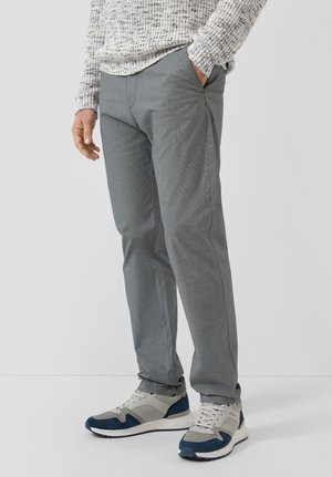 Mann trägt graue Slim-Fit-Hose, weiße und marineblaue Sneakers und einen strukturierten hellgrauen Pullover, mit einer Hand in der Tasche.