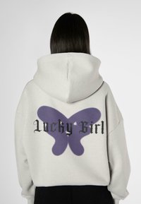 Grå hettegenser med et stort lilla sommerfugl grafikk og teksten "Lucky Girl" i svart på baksiden. Løs passform.