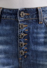 Primer plano de jeans de mezclilla azul con cinco botones metálicos rectangulares decorativos y costuras naranjas visibles en la pretina.