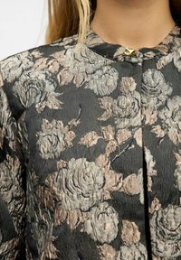Kvinde iført en sort stofjakke med et tekstureret blomsterdesign i beige og grå, der har en lille guldlås ved udskæringen.