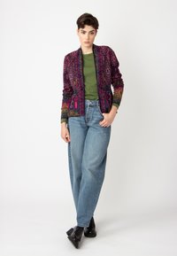 Veste à motifs multicolores avec des teintes violettes, vertes et roses, portée avec un haut vert et un jean taille haute bleu, assortie de chaussures noires.