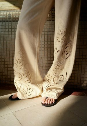 Beige wijde broek met bloemrijke cut-out borduurwerk, gedragen met zwarte open-toe sandalen op een betegelde vloer met zonlicht.