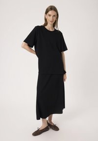 Sort t-shirt med korte ærmer lagd over en sort knælang nederdel. Glat stof med afslappet pasform. Brune flade sko med sløjfer fremhæver outfittet.