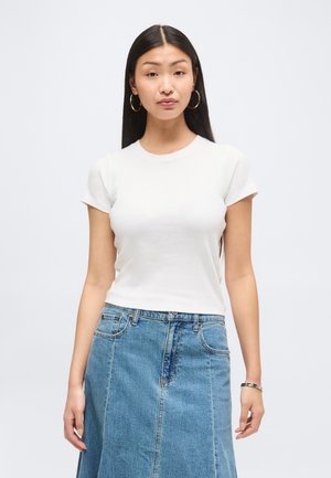 Kvinde med langt sort hår iført guldringe øreringe, hvid tætsiddende top med korte ærmer og blå højtaljet denim nederdel mod ensfarvet baggrund.