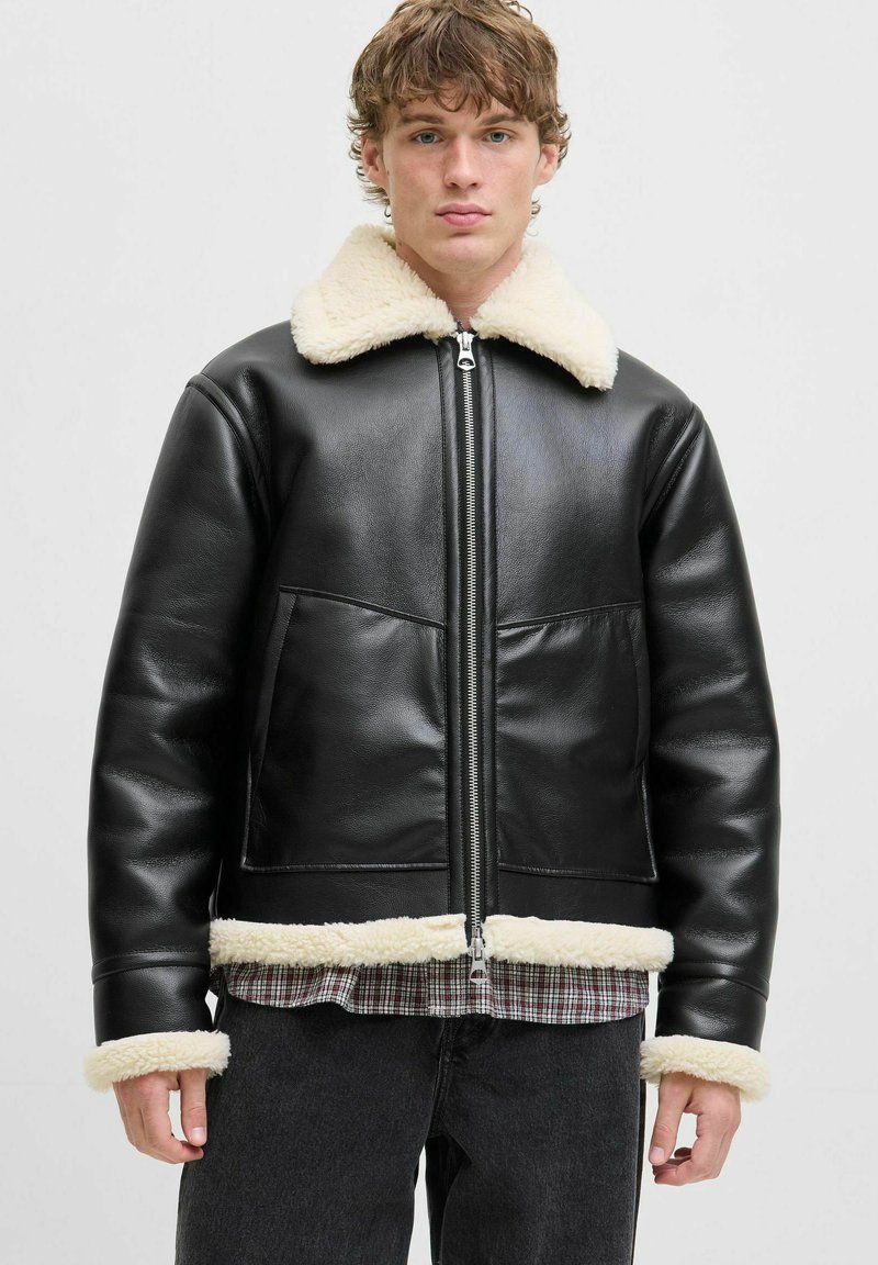 Giovane uomo che indossa una giacca di pelle nera con colletto, polsini e orlo in shearling crema, abbinata a una camicia a quadri e jeans scuri.