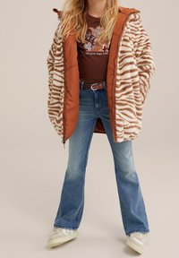 Hnědo-krémový pruhovaný kožich z umělé kožešiny s oranžovými rukávy, kombinovaný s rozšířenými modrými džíny a bílými teniskami.
