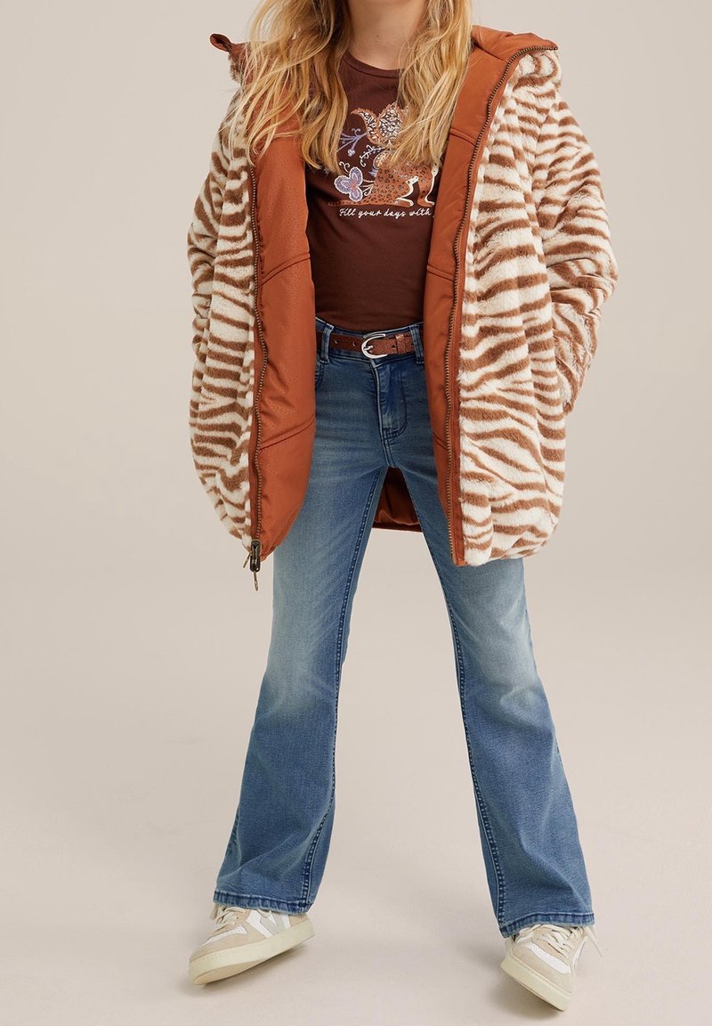 Hnědo-krémový pruhovaný kožich z umělé kožešiny s oranžovými rukávy, kombinovaný s rozšířenými modrými džíny a bílými teniskami.