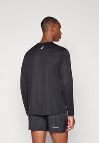 Schwarzes langärmliges Sportshirt mit rundem Ausschnitt und glattem Textil, versehen mit einem weißen Logo hinten, kombiniert mit gemusterten schwarzen Shorts.