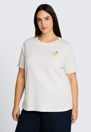 Femme aux longs cheveux foncés portant une chemise blanche à manches courtes avec une petite broderie de citron sur la poitrine gauche, et un pantalon foncé.