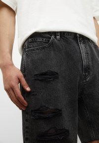 Shorts en jean noir avec des patchs déchirés, coupe standard et design classique à cinq poches. Portés avec un t-shirt blanc basique.
