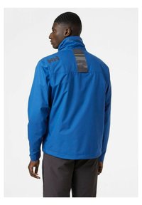 Blauwe waterdichte jas met een hoge kraag, voorzien van een zwart logo op de schouder en een rechthoekig achterpaneel met reflecterende details.