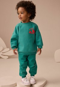 Teal sweatshirt och matchande byxor i mjukt material. Sweatshirten har en röd "BEEP" grafik med en lastbilsdesign. Vita sneakers fulländar outfiten.