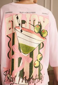 Pink oversized sweatshirt featuring a graphic of a cocktail glass with a straw and olives, accented by colorful stripes and a bow detail.