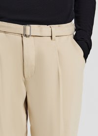 Pantaloni beige su misura con texture liscia, dotati di una fibbia metallica rettangolare e tasche laterali, abbinati a una maglietta nera.
