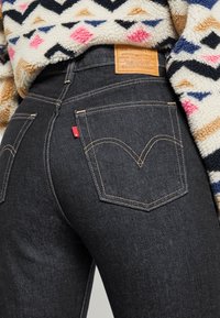 Jean en denim bleu foncé avec coutures jaunes et un patch en cuir marron. Comprend deux poches arrière avec le logo de la marque, partiellement couvertes par une polaire à motifs.