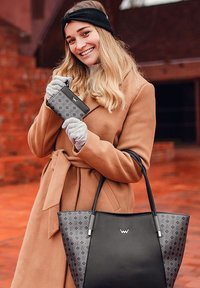 Schwarze und graue gemusterte Tragetasche mit breitem Boden und scharfen Ecken, begleitet von einem kleinen passenden Portemonnaie. Das Modell trägt einen braunen Mantel und Handschuhe.