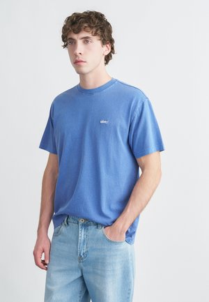 Μπλε βαμβακερό μπλουζάκι με κοντά μανίκια και στρογγυλή λαιμόκοψη, με μικρό λευκό λογότυπο "obey" στο στήθος. Φοριέται με ανοιχτόχρωμα τζιν.