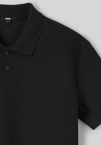 Polo noir en tissu texturé, avec un col classique, une patte de boutonnage à trois boutons et des manches courtes. Étiquette de la marque visible à l'intérieur.