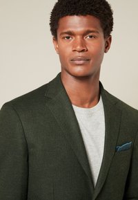 Blazer vert foncé en tissu texturé, coupe unie à simple boutonnage avec revers crantés. Associé à un t-shirt gris clair et un mouchoir de poche bleu.