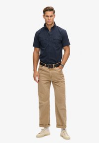 Camisa de manga corta navy con dos bolsillos en el pecho, combinada con pantalones beige claros y zapatillas de crema. Diseño simple, corte recto.