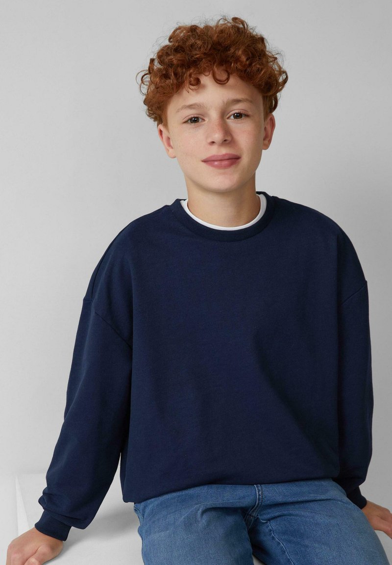 Felpa blu navy con maniche lunghe, scollo rotondo e vestibilità comoda. Indossata sopra una camicia bianca, abbinata a jeans blu, su uno sfondo chiaro.