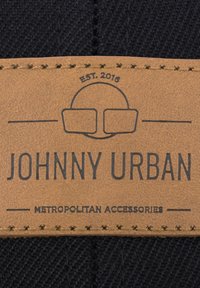 Ruskohvainen nahkaleima, jossa on kohokuvioitu teksti "JOHNNY URBAN" tumman kudotun tekstiilin taustalla. Sisältää siistiä ompelua ja suunnittelu elementtejä.