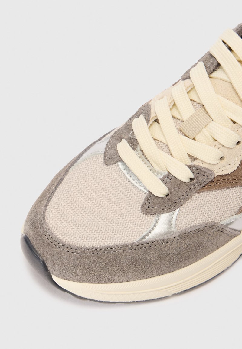 Beige und graue Sneaker aus strukturierten Stoffen und Wildleder. Mit cremefarbenen Schnürsenkeln und metallischen Akzenten an der Oberseite. Runde Zehenform und dicke Sohle.