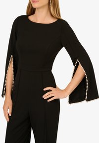 Černý overal s rozšířenými rukávy, zdobený perlovými akcenty podél švů. Hladký materiál s těsným tělem a širokými nohavicemi.