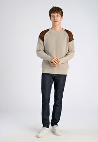 Vaahto beige ribbed-sweateri, jossa on ruskeat olkaimet, pyöreä kaula-aukko ja pitkät hihat. Yhdistetty tummien farkkujen ja valkoisten lenkkarien kanssa.
