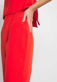 Tenue en tissu rouge comprenant un haut ample et un pantalon ajusté. Le matériau semble lisse avec un léger éclat et des poches latérales sur le pantalon.