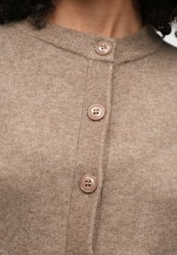 Beige knitted cardigan with a round neckline, featuring four light brown buttons and a soft texture, highlighting its simple design and functionality.