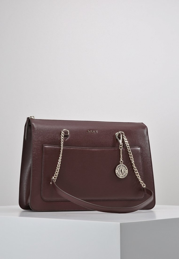 Burgundská kožená kabelka s přední kapsou, stříbrnými řetízkovými uchy a přívěskem s logem. Hladká textura s propracovaným designem.