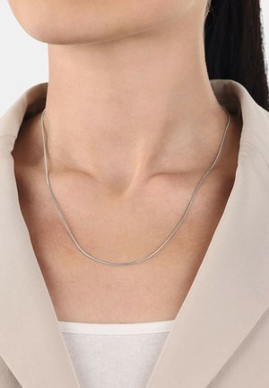 Collar de plata con un diseño liso y curvado, reposando sobre un cuello. El fondo es neutro, con un blazer beige claro visible.