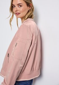 Veste bomber rose avec une texture lisse, ourlet côtelé et poches latérales. Comprend une poche zippée sur la manche gauche et une coupe décontractée.