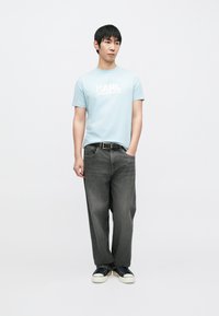 Világoskék pamut póló "KARL LAGERFELD" logóval, laza szabású sötétszürke farmernadrággal és fekete, fehér talpú tornacipővel párosítva.