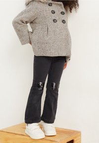 Grå texturerad kappa med svarta knappar, i kombination med svarta jeans som har paljetthjärtan på knäna, och vita sneakers.