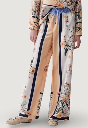 Szerokie spodnie z satyny z pionowymi granatowymi paskami i kwiatowymi wzorami w kolorze brzoskwiniowym i białym, zestawione z dopasowaną kwiatową bluzką i beżowymi sandałami z ozdobnymi kamieniami.