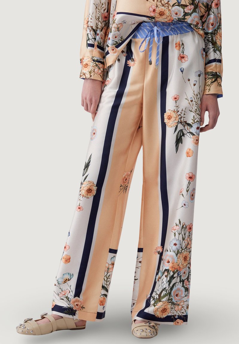 Weit geschnittene Satinhosen mit vertikalen marineblauen Streifen und Blumenmustern in Pfirsich und Weiß, kombiniert mit einem passenden floralen Oberteil und beigen Sandalen mit Schmucksteinen.