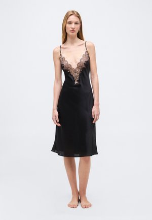 Woman standing barefoot wearing a black slip dress with intricate lace detailing on the deep V neckline against a plain light background.