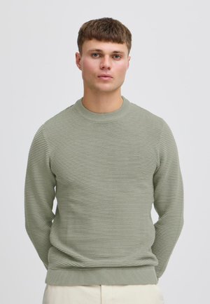 Młody mężczyzna o krótkich brązowych włosach, ubrany w jasnozielony prążkowany sweter i beżowe spodnie, stojący na tle jednolitego, jasnego tła.