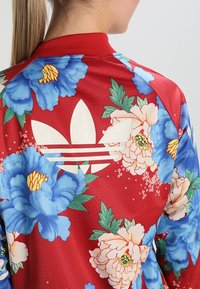 Röd blommig bomberjacka med blå pioner och vita pionmönster. Den har en vit adidas-logotyp och en ribbad krage.