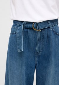 Denim trousers with a high waist, light blue color, wide leg, and a fabric belt featuring a gold circular buckle. Central pleats add detail.