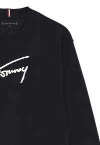 Maglione nero girocollo con ricamo corsivo bianco "Tommy" sul petto e una piccola etichetta Tommy Hilfiger all'interno del colletto.