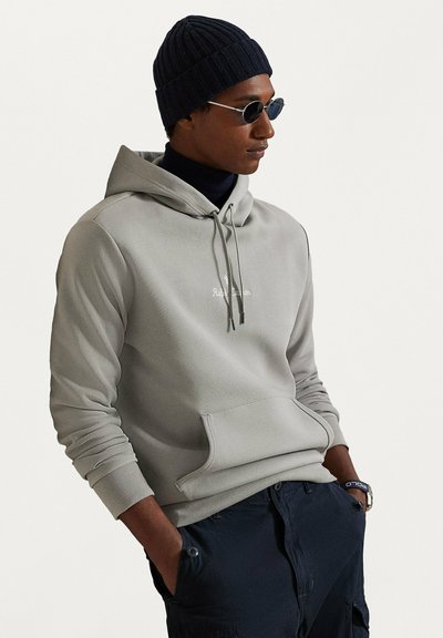 Felpa con cappuccio grigio chiaro con tasca a marsupio, cappuccio con coulisse e logo ricamato. Abbinata a un berretto a coste blu navy e occhiali da sole tondi.