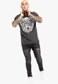 Schwarzes Grafik-T-Shirt mit einem zentralen weißen Totenkopfdesign und abgewetzten schwarzen Jeans mit aufgerissenen Knien, kombiniert mit weißen Turnschuhen.