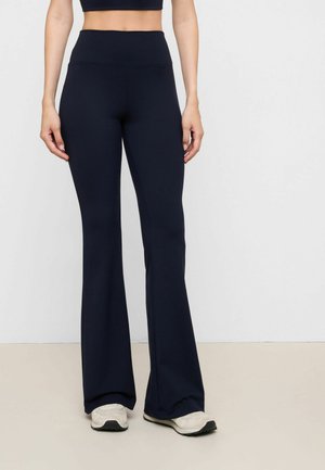 Kvinde iført sorte højtaljede, vide leggings og hvide sneakers, stående mod en ensfarvet hvid baggrund.