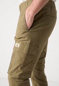 Olivgrön cargo-byxor med en strukturerad tyg, som har en sidoficka och en logotypapplikation, tapered ben-design och elastisk midja.